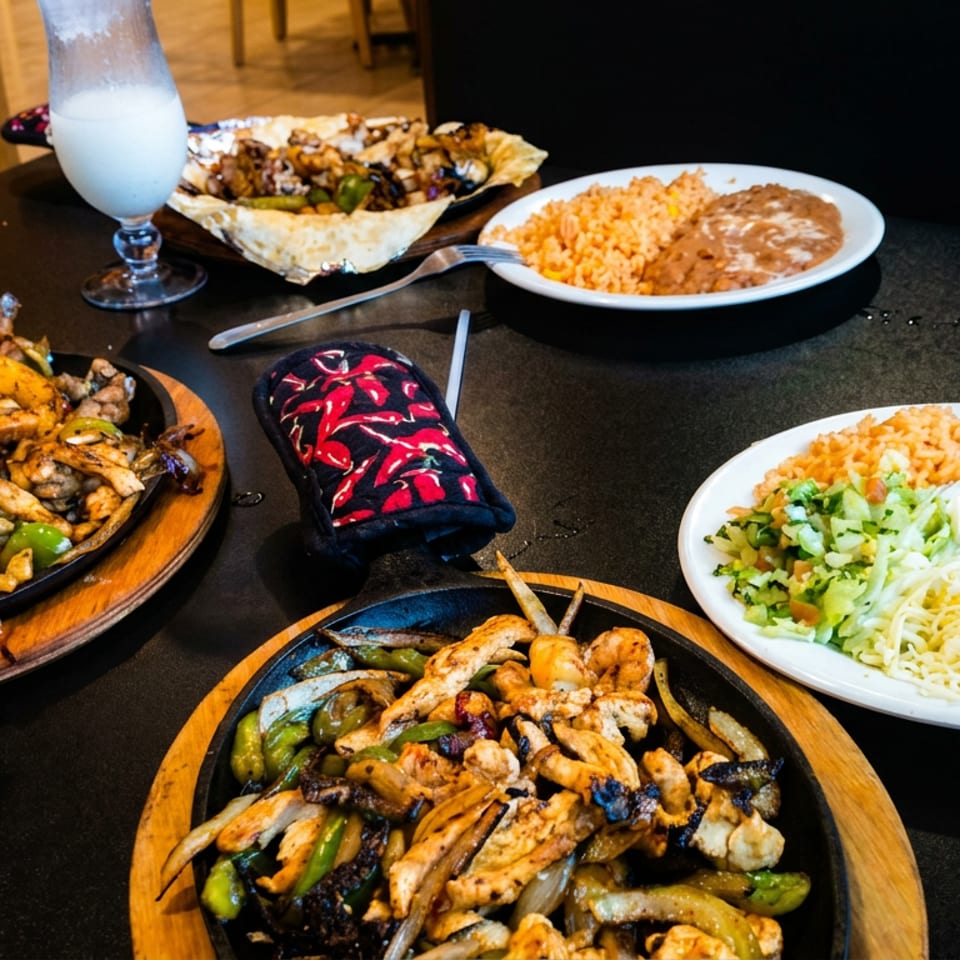 Mexican restaurant table featuring sizzling fajita platters, rice and beans, guacamole, chips, and colorful beverages.